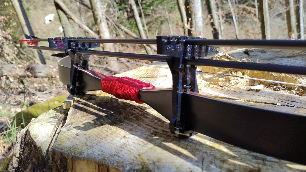 Nahaufnahme eines Bogens mit einer Pfeilhalterung, die auf einem Baumstumpf platziert ist. Der Bogen hat eine rote Schnur um den Griff und Pfeile sind auf der Halterung befestigt, mit natürlicher Waldumgebung im Hintergrund.