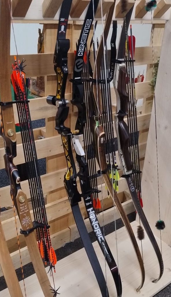 Eine Reihe von verschiedenen Bögen und Pfeilen, die an einem Holzregal ausgestellt sind. Bogenköcher Q7