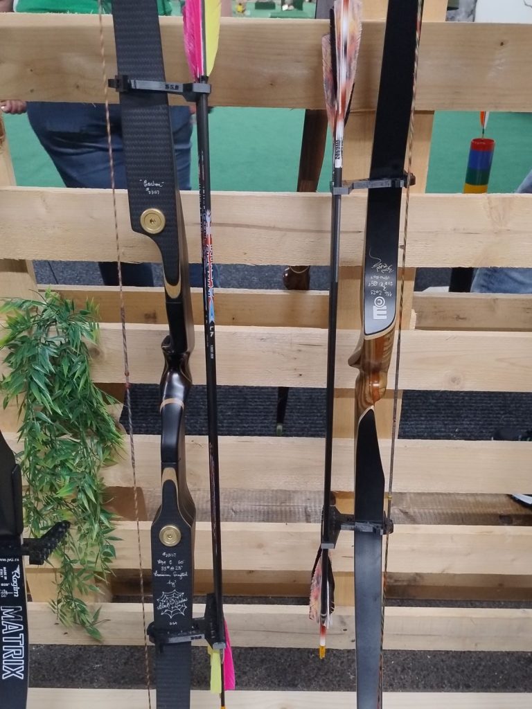 Verschiedene Bögen mit Pfeilen in einem Holzregal, ausgestattet mit einem Bogenköcher-System zur einfachen Aufbewahrung und Organisation der Pfeile. Bogenköcher Q7